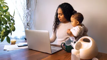 A young mixed race African American mother holds her daughter while taking notes at her dining table serving as a temporary remote work from home station with breast pump in foreground.